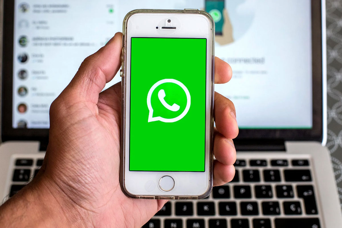 Una persona tiene in mano un cellulare con la schermata iniziale di WhatsApp, sullo sfondo un computer